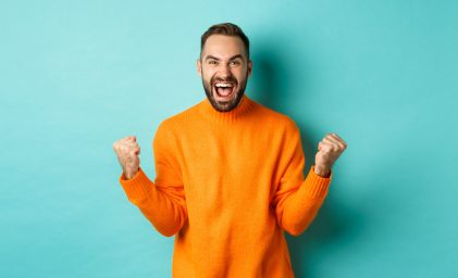 Excited man celebrating victory, rejoicing and making fist pump gesture, winning and looking satisfied, saying yes, achieve goal, standing over light blue background.